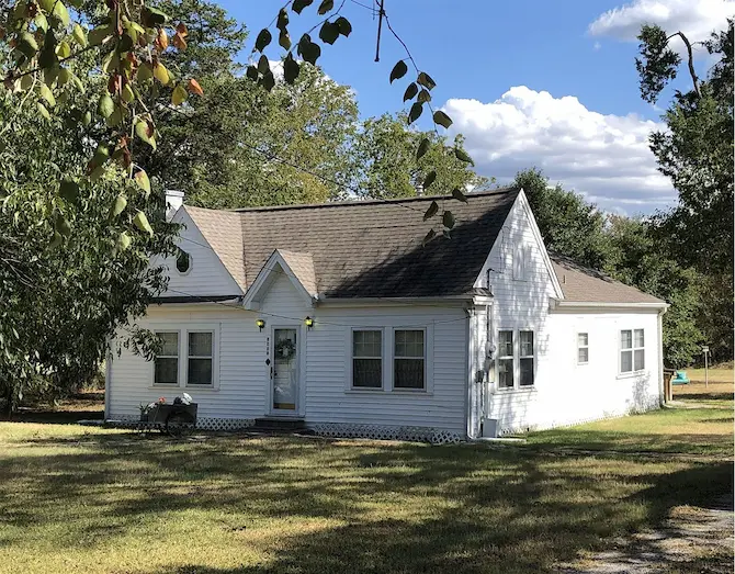 A charming small white house with a welcoming porch, surrounded by lush green trees, perfect for a cozy family home.