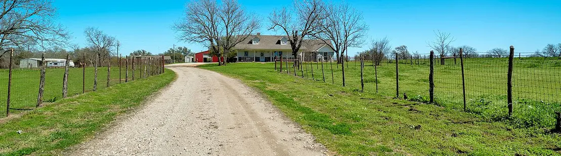 A dirt road winds through greenery, leading to a charming farmhouse surrounded by open fields.