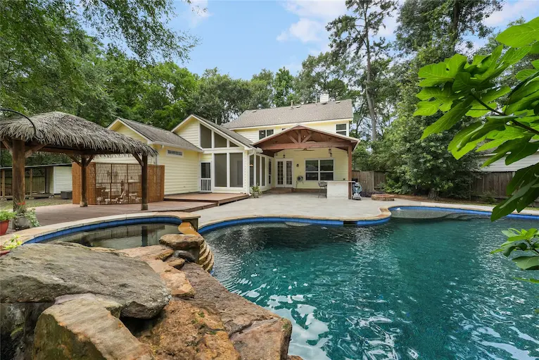 A serene pool and patio area featuring a gazebo, surrounded by a well-maintained landscape and a charming house nearby.